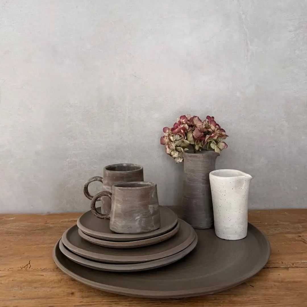 AREIA Natural Geschirr-Set mit Tellern, Bechern und kleine Krüge/Vasen/Kännchen, auf Holztisch vor grauem Hintergrund