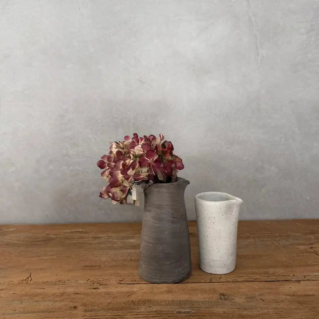 AREIA Natural Kännchen/Vase mit Trockenblumen neben hellem Keramikbecher, auf Holztisch vor grauem Hintergrund