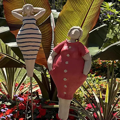 Zwei Keramikfiguren – Badedame mit blau-weißem Streifenkleid und Frau im roten Knopfkleid, zwischen Bananenblättern