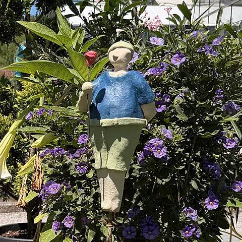 Keramikfigur eines Mannes in Blau mit grüner Hose und mit roter Blume, zwischen violetten Blüten