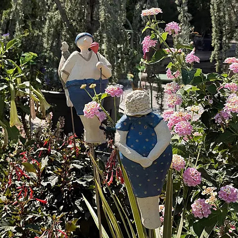 Zwei Keramikfiguren zwischen Lantana-Blüten – Mann in Blau mit Blume und Frau in blau-gepunktetem Kleid mit Hut