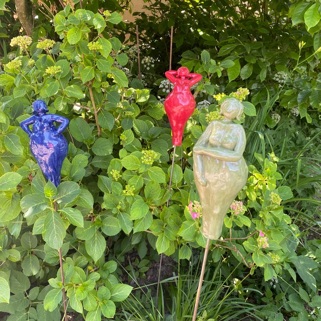 Drei Keramikfiguren auf einem Stab. Zur Dekoration vor einem Strauch. Mittelblau-, apfelgrün- und rotglänzend.