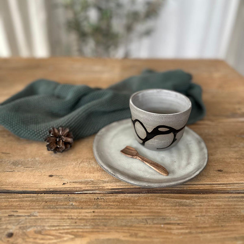 Nahaufnahme des Keramikbechers mit schwarzer Schlangenlinie und weiß glasiertem Inneren sowie mit weiß glasiertem Teller, handgefertigtes Unikat