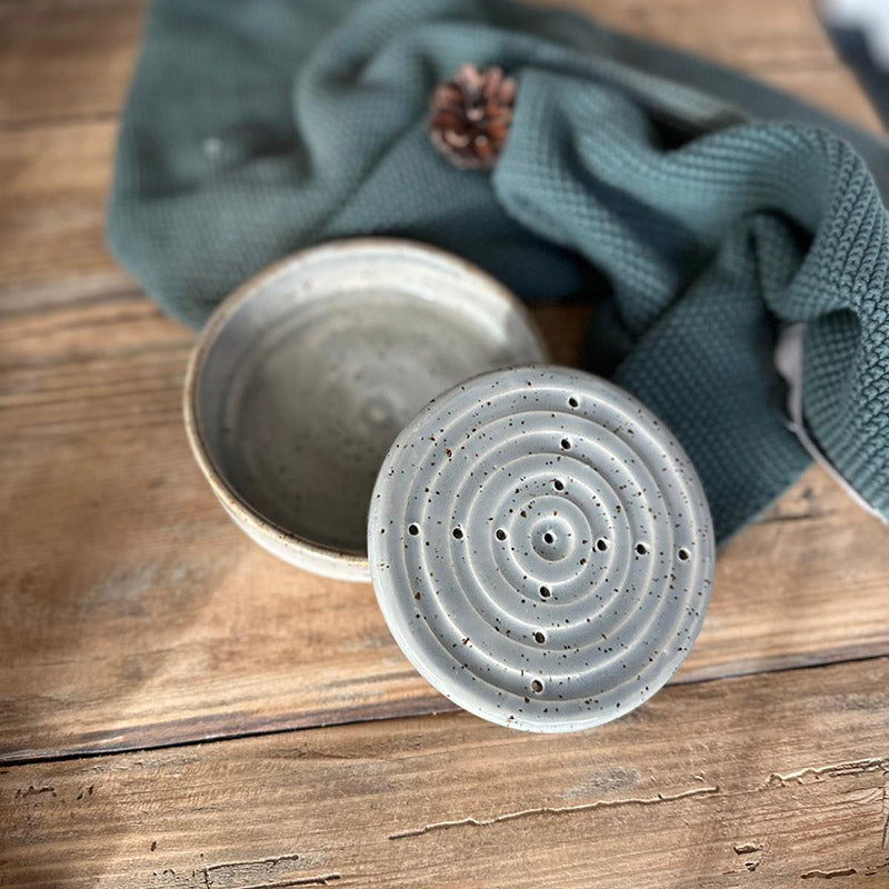 Runde graue Seifenschale mit Spots, dezente Bad-Deko neben strukturiertem Tuch, Oberschale liegt schräg auf Unterschale