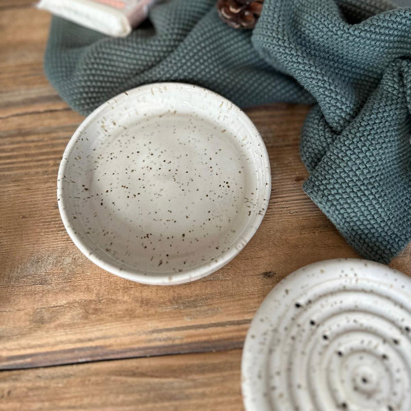 Minimalistische weiße Steinzeug-Seifenschale mit Spots neben grünem Waffeltuch, beide Schalen liegen nebeneinander