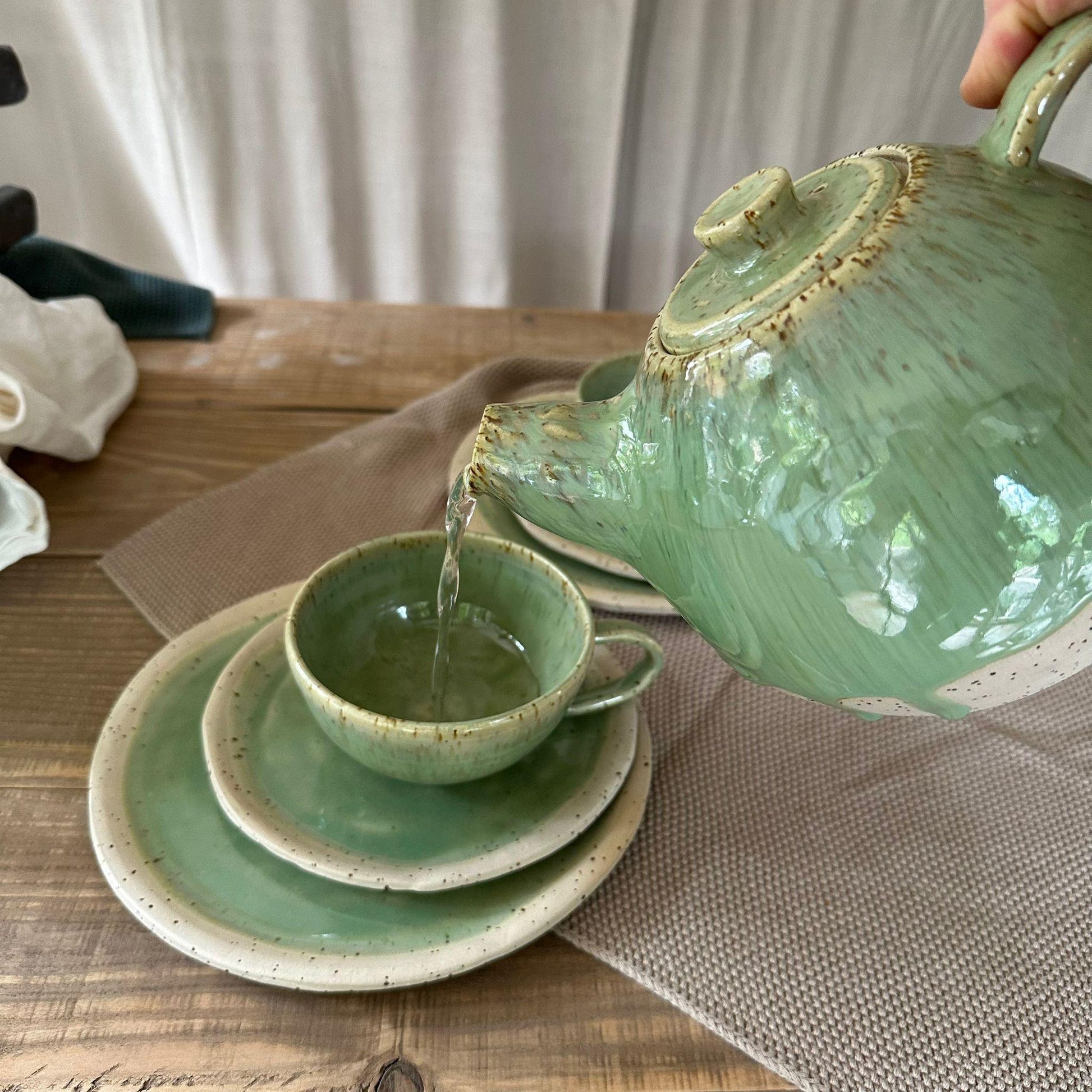 Tee Geschirr Set aus Steinzeugton mit Spots. Grüne, glänzende Glasur. Aus der bauchigen Teekann wird Tee in eine Teetasse gegossen.